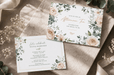 A styled flat-lay of a floral afternoon tea invitation card with gold foil stamped text, displayed on a dark surface alongside a matching RSVP card, envelope, and dried baby's breath flowers, printed by Clubcard Palm Springs on 12pt 100% recycled fibers stock with full color detail.