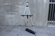 Folding aluminum easel stand with transportation bag on a grey concrete background.